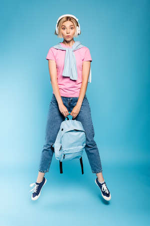 blonde student in wireless headphones looking at camera while holding backpack and jumping on blue
