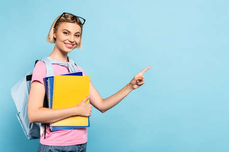 young blonde student with backpack holding notebooks and ponging with finger on blue