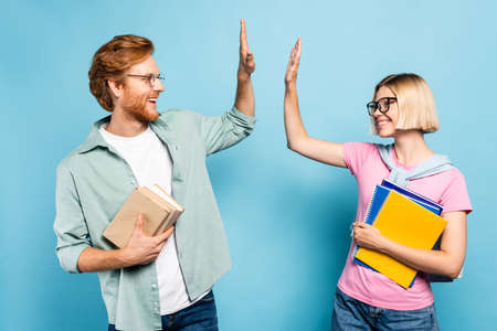 young students in glasses holding notebooks and books while giving high five on blue
