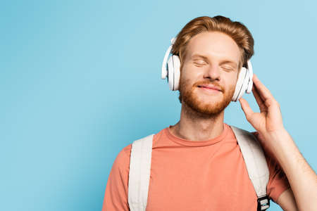bearded and redhead student with closed eyes touching wireless headphones on blueの写真素材