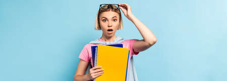 panoramic shot of shocked student touching glasses while holding notebooks on blue