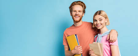 panoramic shot of bearded man holding notebooks and hugging blonde student with books on blue