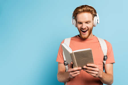 excited student in wireless headphones reading book on blue