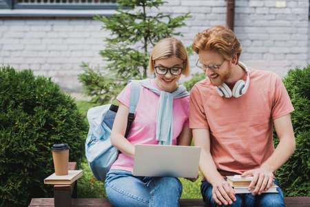 students in glasses looking at laptop while sitting on bench near books and paper cupの写真素材