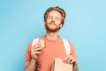 young redhead student with closed eyes holding paper bag and toast bread on blueの写真素材