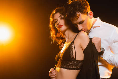 passionate man in suit taking off jacket on sexy woman in bra on black with back lightの写真素材