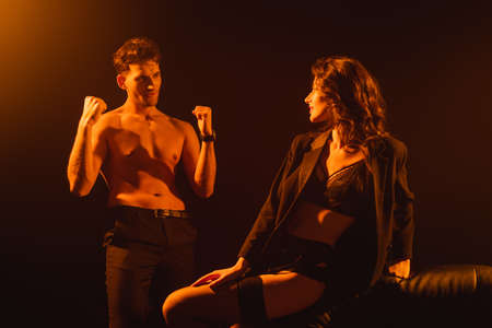 seductive woman in jacket looking at shirtless man on black