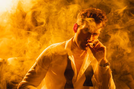 man in suit holding cigar and looking at camera on black with smokeの写真素材