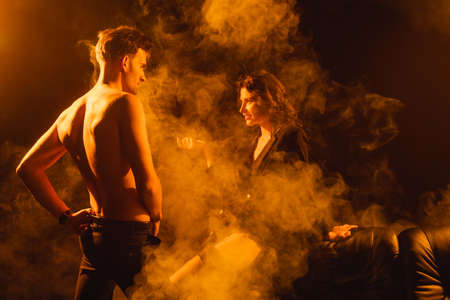 curly woman in jacket looking at shirtless man standing with hand on hip on black with smoke