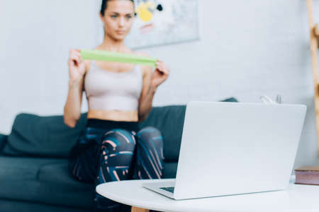 Selective focus of laptop on coffee table and sportswoman holding resistance band on couchの写真素材
