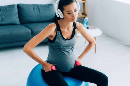 Exhausted pregnant sportswoman in headphones training with dumbbells on fitness ball at homeの写真素材