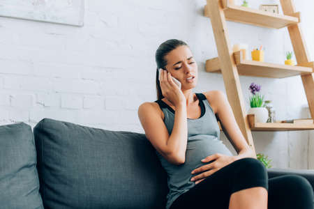 Selective focus of pregnant sportswoman taking on smartphone while feeling pain on couchの写真素材