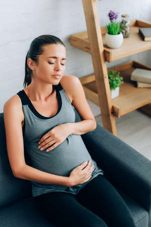 Pregnant sportswoman feeling pain and embracing belly on couchの写真素材