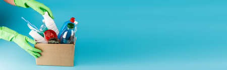 cropped view of cleaner in rubber gloves collecting rubbish in cardboard box on blue background,...