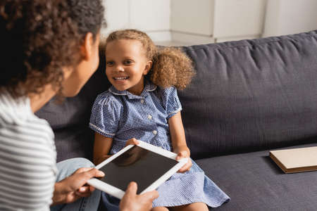 selective focus of nanny holding digital tablet with blank screen near african american girl sitting...