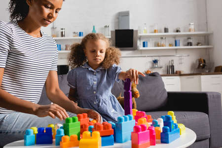 african american girl in dress and babysitter in striped t-shirt playing with colorful building blocks in kitchenの写真素材