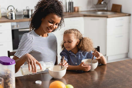 african american mom in striped t-shirt pouring milk into bowl while having breakfast with daughterの写真素材