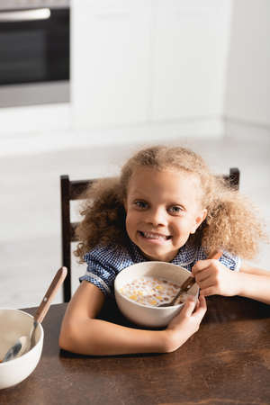high angle view of excited african american girl looking at camera while holding spoon near bowl...