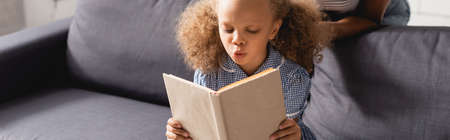 cropped view of nanny behind focused african american girl sitting on sofa and reading book, website...