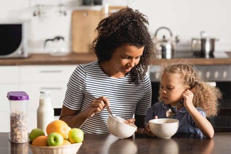 african american woman in striped t-shirt showing bowl with breakfast to daughterの写真素材