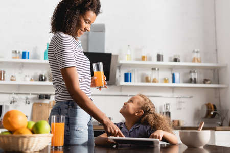 young african american woman holding orange juice and touching digital tablet near daughter in kitchenの写真素材
