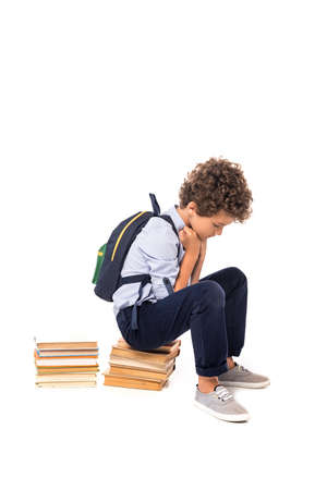 upset schoolboy with backpack sitting on books isolated on whiteの写真素材