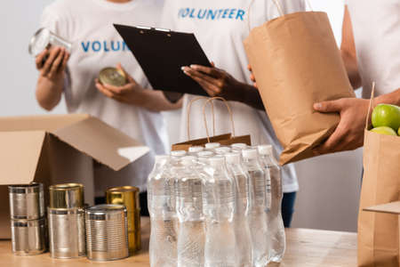 Cropped view of multiethnic volunteers with clipboard standing near bottles of water and packages...