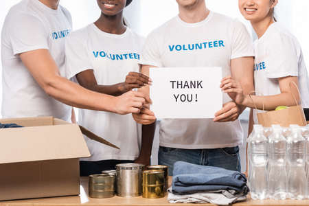 Cropped view of multiethnic volunteers holding card with thank you lettering and donations in...