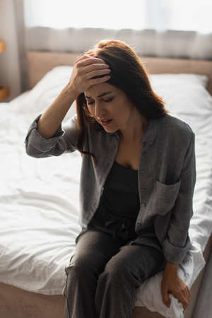 tired woman with closed eyes suffering from pain and touching head while sitting on bed at homeの写真素材