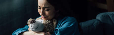panoramic crop of sad brunette woman with closed eyes holding teddy bear in living room