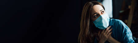 panoramic crop of scared brunette woman in blue medical mask touching throat at homeの写真素材