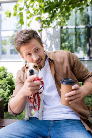 young man with jack russell terrier dog holding coffee to go and pointing with finger while sitting on benchの写真素材