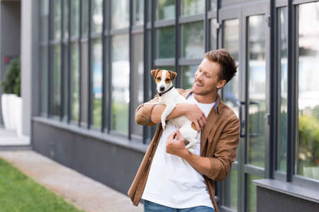 young man in casual clothes holding jack russell terrier dog near building with glass facadeの写真素材