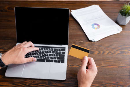 Cropped view of businessman holding credit card and using laptop with blank screen in office