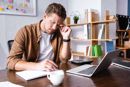 Selective focus of pensive businessman writing on notebook beside cup of coffee, laptop and...