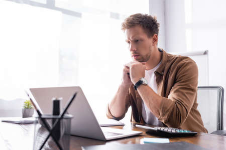 Selective focus of young businessman looking at laptop near calculator and stationery in officeの写真素材