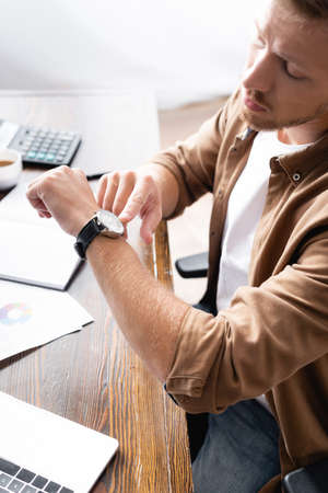 Selective focus of businessman pointing at wristwatch while working with laptop and papersの写真素材