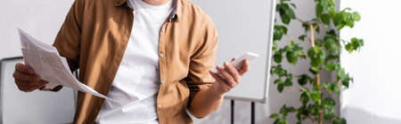 Panoramic crop of businessman holding papers and smartphone in office