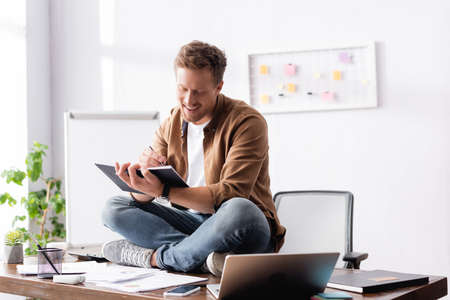 Selective focus of young businessman writing on notebook while sitting beside gadgets and documents...