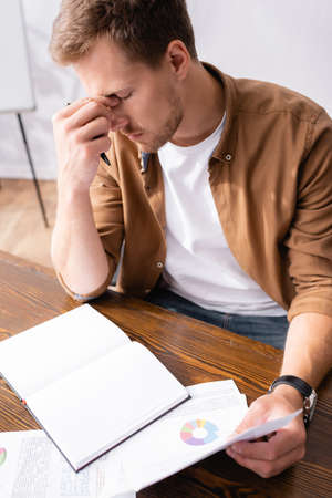 Selective focus of tired businessman holding papers near notebook on table in officeの写真素材