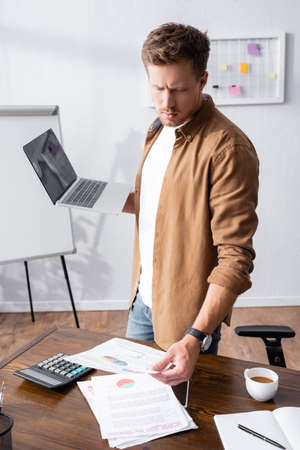 Selective focus of young businessman looking at paperwork and holding laptop near calculator and coffee in officeの写真素材