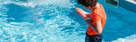 panoramic crop of boy playing with water gun near pool