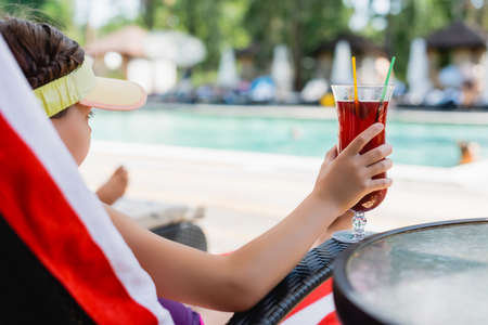 selective focus of girl in sun visor cap holding cocktail glass while resting in deck chairの写真素材