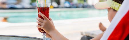 horizontal image of girl in sun visor cap holding cocktail glass while resting in deck chair