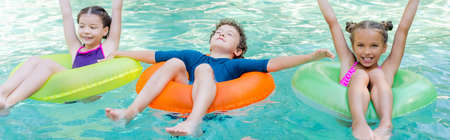 panoramic concept of girls with hands in air and boy with closed eyes swimming in pool on inflatable...