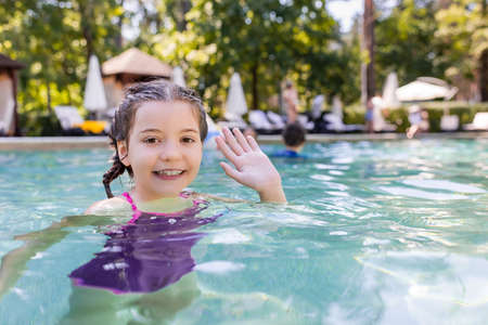 joyful girl in swimming pool looking at camera and waving handの写真素材