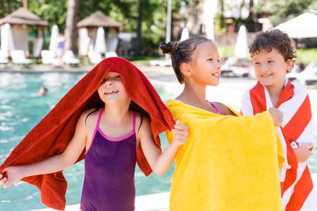 excited girl in swimsuit covering head with towel while standing near pool with friendsの写真素材