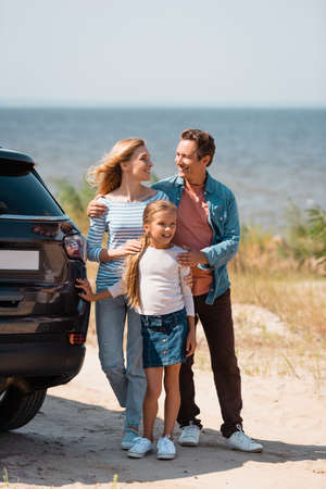 Selective focus of man and woman hugging daughter near car on seasideの写真素材