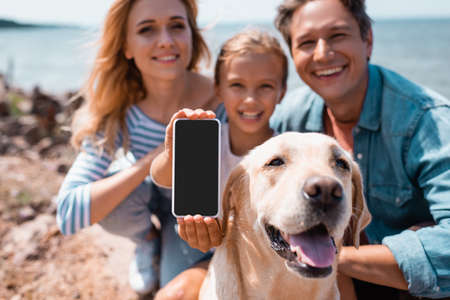 Selective focus of woman holding smartphone near family and golden retriever on seasideの写真素材