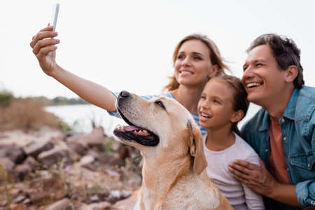 Selective focus of family with golden retriever taking selfie on smartphone on beachの写真素材